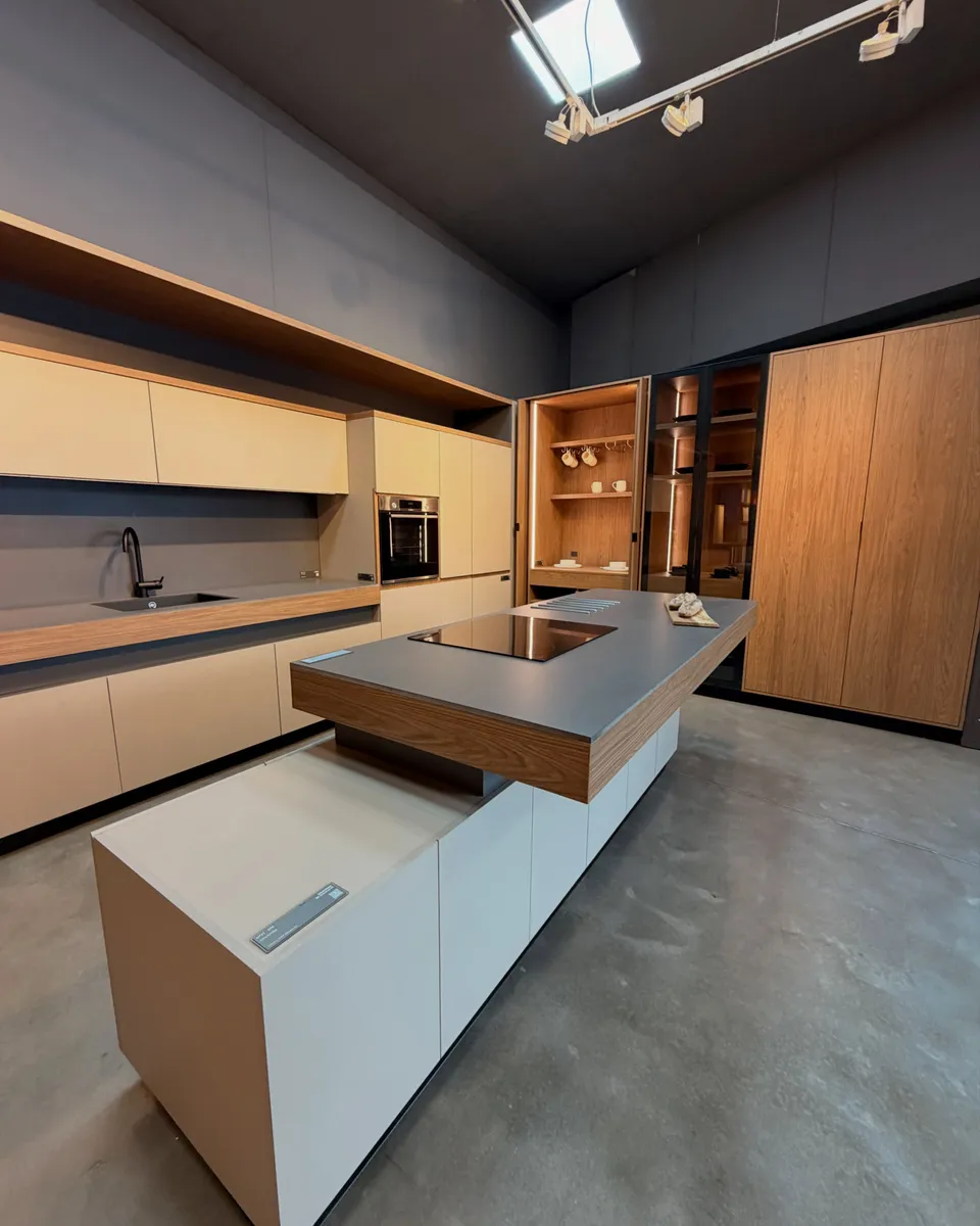 Cocina completa en el showroom, con isla central, frentes en melamina clara y paneles de madera.